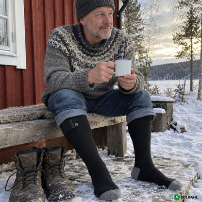 Man utanför röd stuga i snön med värmestrumpor och en kopp kaffe